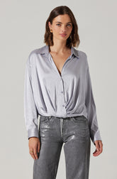 A woman is wearing a light gray satin button-up shirt with a subtle dotted pattern. The shirt has a classic collar, long sleeves with cuffs, and is slightly oversized, with the front loosely gathered at the waist. She is also wearing high-waisted, gray washed jeans.